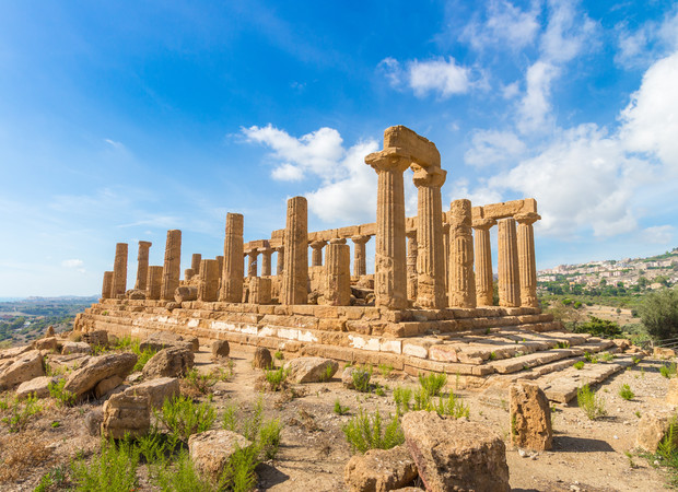 Ancient stone temple with tall, weathered columns stands under a bright blue sky, surrounded by scattered stones and sparse green vegetation in a dry, open landscape.